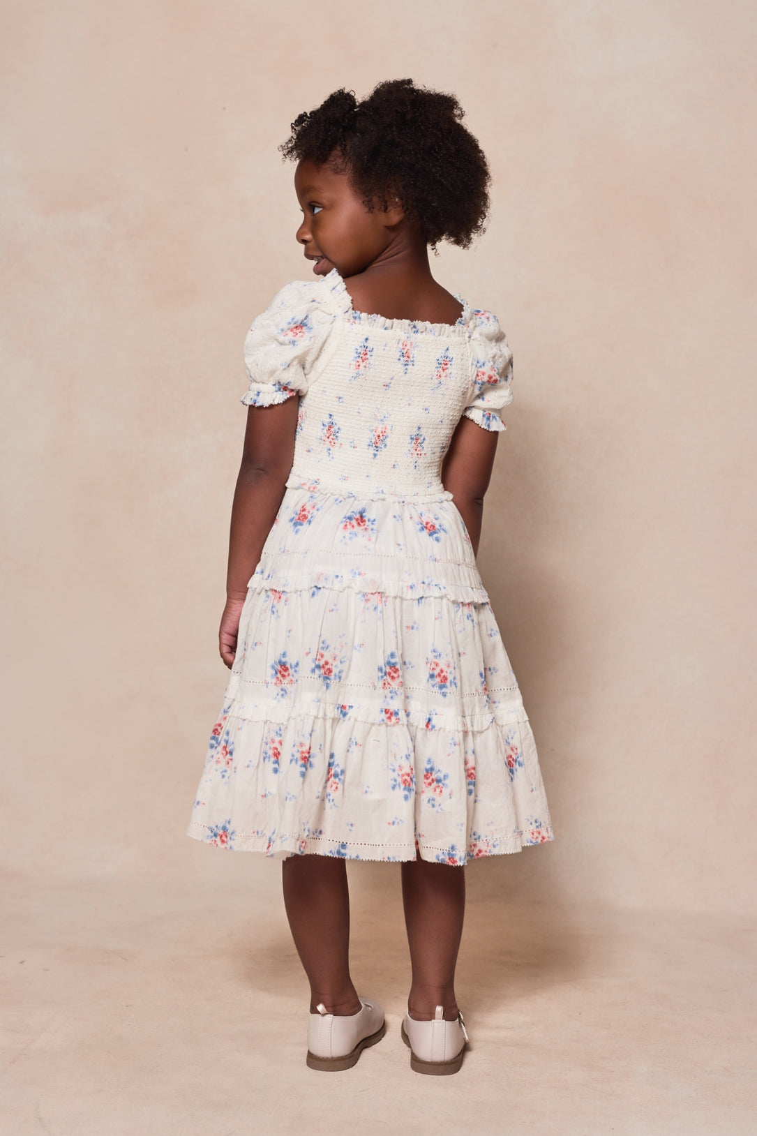 A young girl with curly hair faces away from the camera on a beige background, wearing the Ivy City Co Mini Emily Dress in Heritage Soft Ditsy Floral and light-colored shoes.