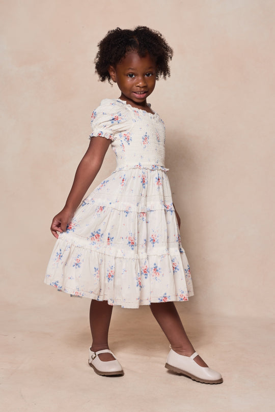 A young girl with curly hair and white shoes smiles as she poses in Ivy City Co's Mini Emily Dress in Heritage Soft Ditsy Floral, standing against a neutral background.