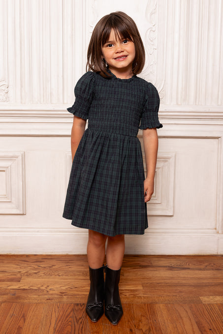 Young girl wearing a dark plaid dress standing against a white paneled wall.