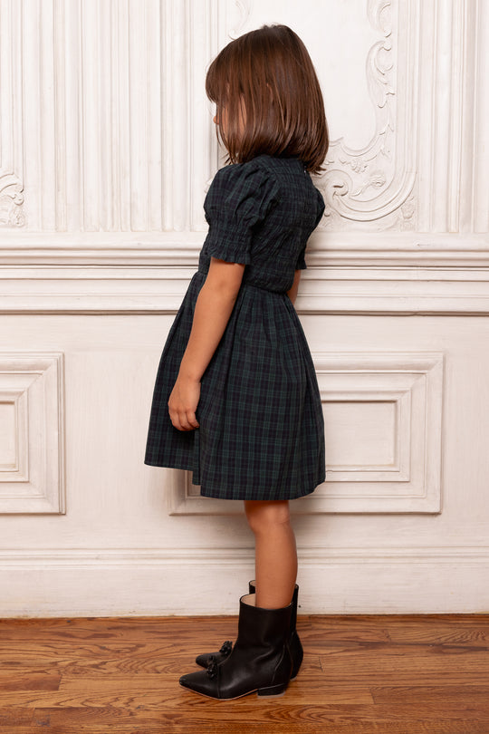 Child wearing a dark plaid dress and black shoes against a decorative white wall.