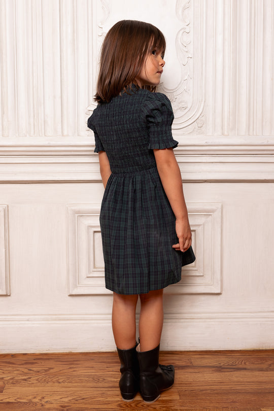 Young girl wearing a checkered dress standing against a white wall.