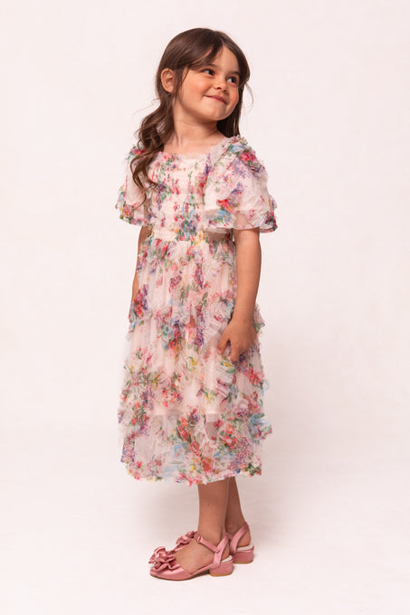 A young girl with long brown hair wears the Ivy City Co Mini Florentina Dress in Pink Floral and pink bow sandals, standing and smiling while looking slightly left against a light background.