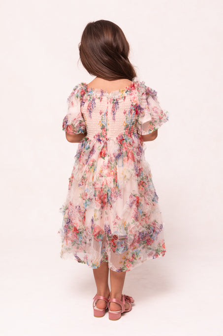 A young girl with long brown hair stands facing away, wearing the Ivy City Co Mini Florentina Dress in Pink Floral with puffed sleeves and pink sandals, set against a plain light background.