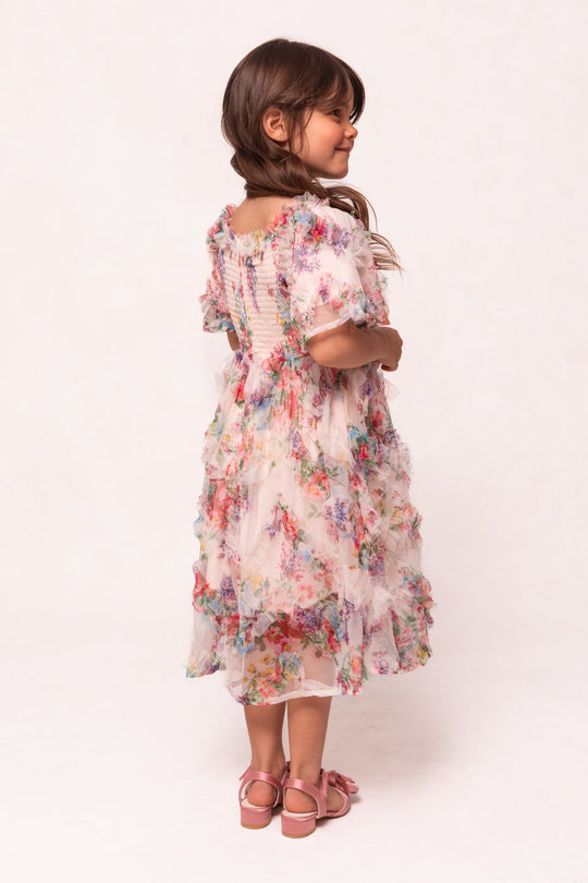 A young girl with long brown hair in pigtails wears the Mini Florentina Dress in Pink Floral by Ivy City Co, styled with pink sandals. She stands sideways, looking left, against a light background.