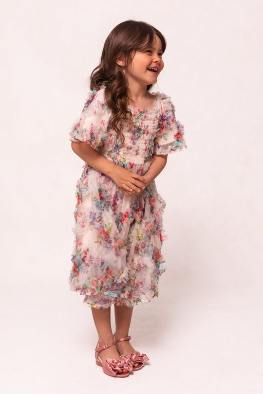 A young girl with long brown hair smiles in the Ivy City Co Mini Florentina Dress in Pink Floral, a girls’ midi dress with ruffled short sleeves, paired with pink bow sandals against a light background.
