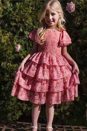A young girl with long blonde hair smiles outdoors, standing in front of green bushes and pink roses. She wears the Ivy City Co Mini Grace Dress in Desert Heritage Rose, a pink floral ruffled flutter sleeve dress, and holds the sides of her dress.