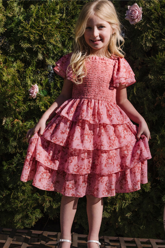 A young girl with long blonde hair smiles outdoors, standing in front of green bushes and pink roses. She wears the Ivy City Co Mini Grace Dress in Desert Heritage Rose, a pink floral ruffled flutter sleeve dress, and holds the sides of her dress.