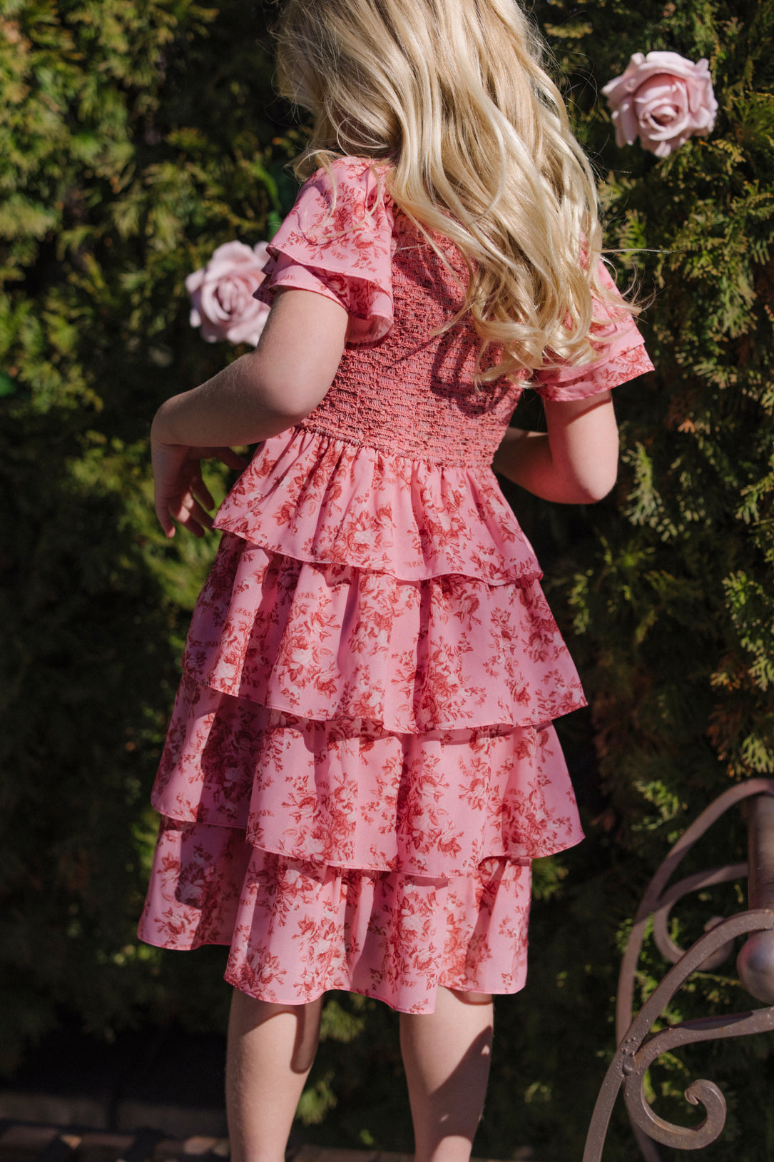 A young girl with long blonde hair wears the Ivy City Co Mini Grace Dress in Desert Heritage Rose with ruffled details, standing outdoors near green shrubs and blooming pink roses, facing away from the camera.