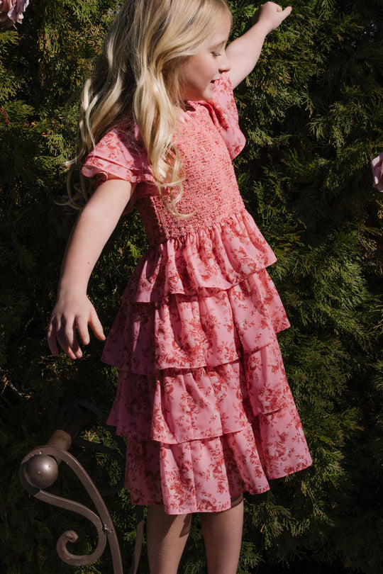 A young child with long blonde hair wears the Mini Grace Dress in Desert Heritage Rose by Ivy City Co, a pink floral maxi dress with ruffles, standing outdoors with arms outstretched before green shrubbery.