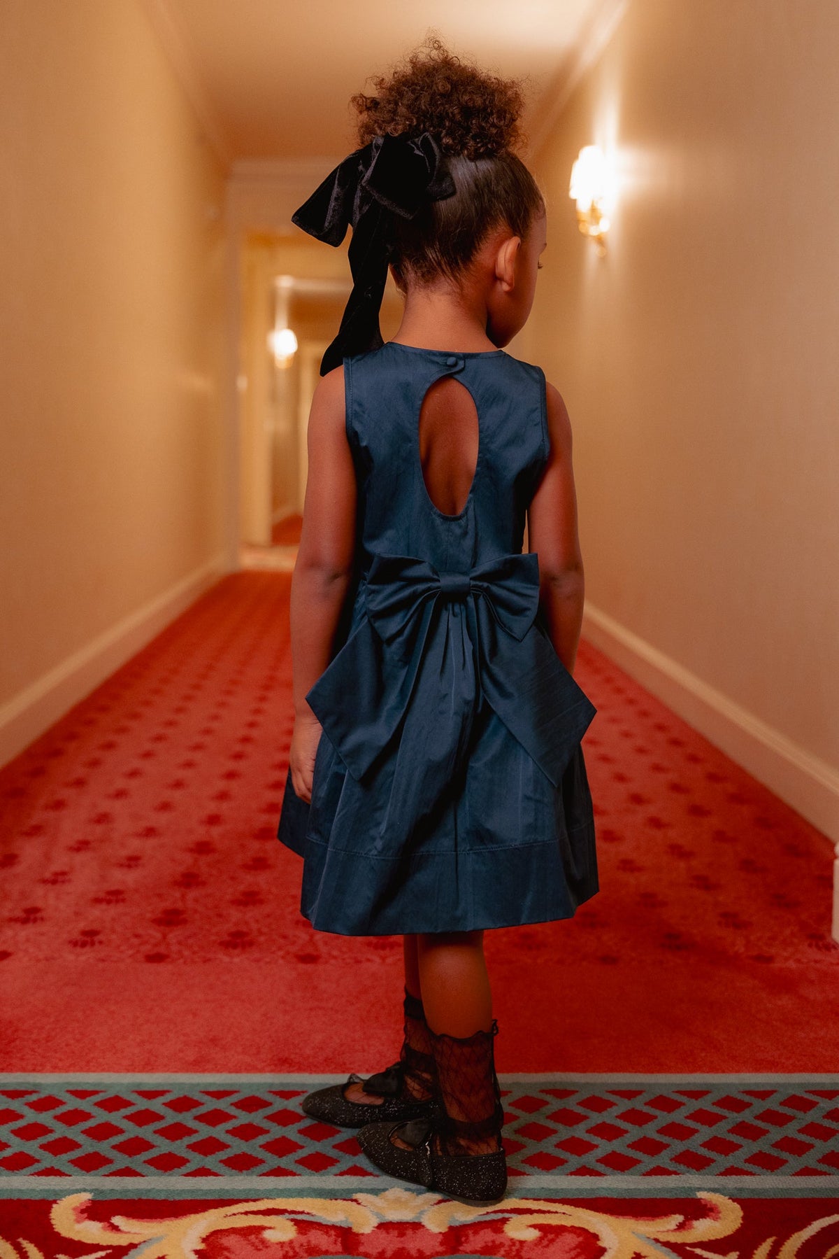 A young girl wearing the Ivy City Co Mini Greyson Dress in Navy, distinguished by its large bow detail, stands on a red carpet in a well-lit hallway with her back to the camera.