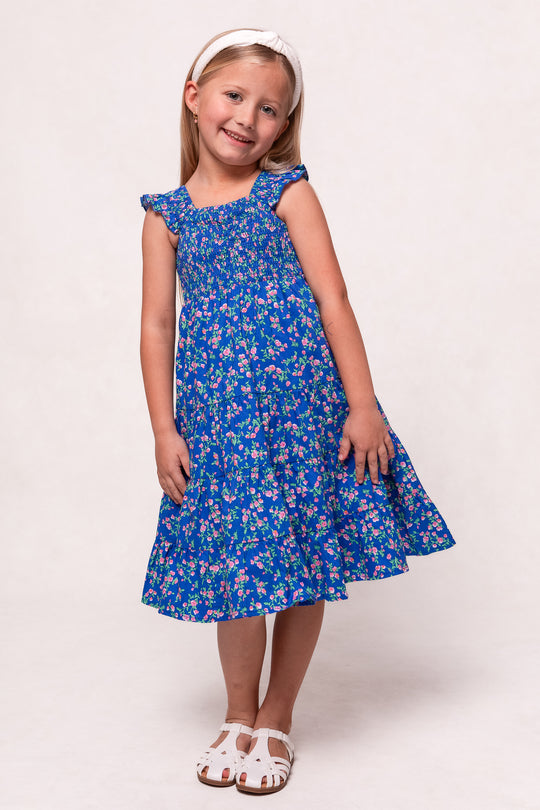A young girl smiles in the Mini Hailey Dress in Allie Blue Floral by Ivy City Co—a blue floral cotton midi with ruffled sleeves and hem—paired with white sandals and a headband, standing against a light background.