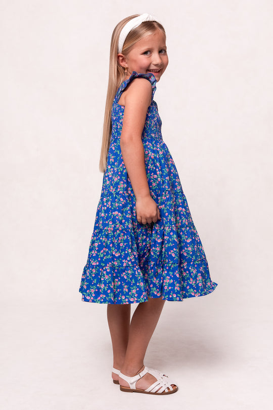 A young girl in the Ivy City Co Mini Hailey Dress in Allie Blue Floral, paired with a white headband and sandals, stands sideways and smiles softly against a plain light background.