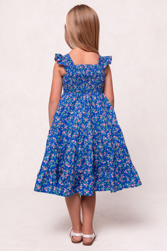 A young girl with long blonde hair wears the Ivy City Co Mini Hailey Dress in Allie Blue Floral—a blue floral cotton midi dress with ruffled sleeves—and white sandals, standing with her back to the camera against a light background.