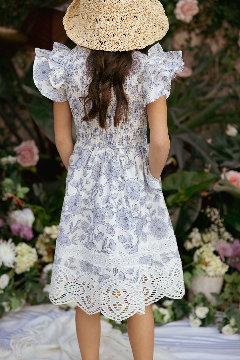 A young girl wearing the Ivy City Co Mini Hattie Dress in Blue Floral and a wide-brimmed straw hat stands outdoors, facing away, surrounded by flowers and greenery.