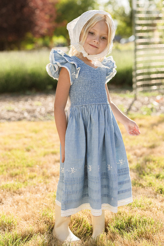 A young child stands outdoors on the grass with one hand in her pocket, wearing the Ivy City Co Mini Hattie Dress in Embroidered Chambray with flutter sleeves, paired with a white bonnet and boots. She looks at the camera, surrounded by sunlight and greenery.