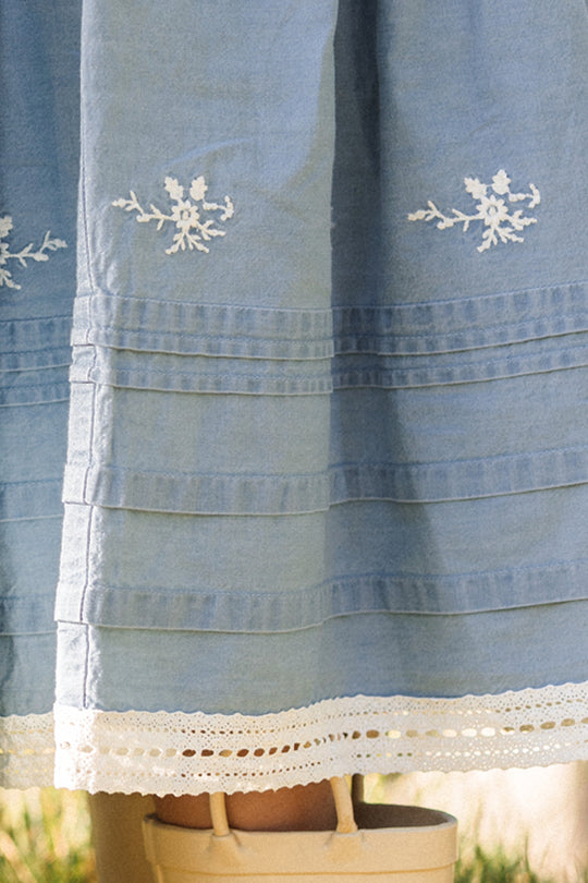 A close-up of the Ivy City Co Mini Hattie Dress in Embroidered Chambray showcases its light blue fabric adorned with white floral embroidery, horizontal pleats, and delicate lace trim at the hem—signature details of this style. The lower part of a person’s legs and their beige shoes appear partially at the bottom of the image.