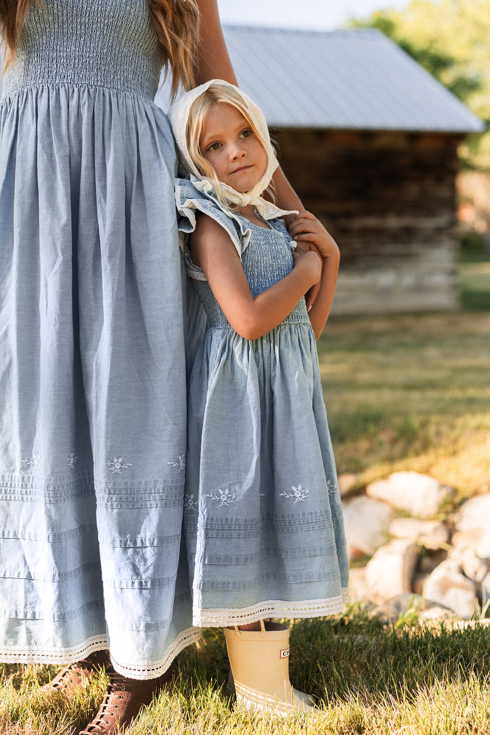 A young girl wearing the Ivy City Co Mini Hattie Dress in Embroidered Chambray, featuring a smocked bodice and flutter sleeves, stands outdoors beside a woman in the matching dress near a rustic wooden building, with grass and rocks in the background.