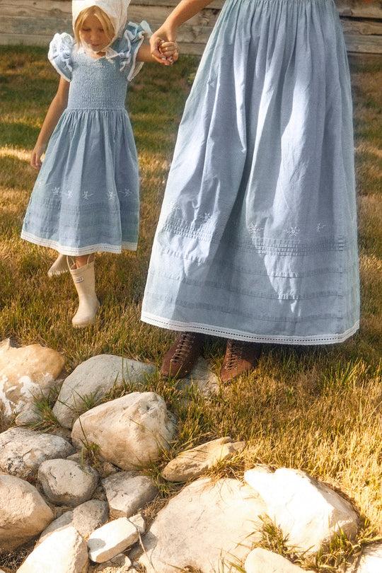 A woman and young girl, both wearing Ivy City Co’s Mini Hattie Dress in Embroidered Chambray—with smocked bodices and light blue fabric—hold hands as they walk on grass near smooth rocks. The woman pairs her dress with brown lace-up boots, while the girl wears white shoes and a matching white headscarf. Their faces are not shown.