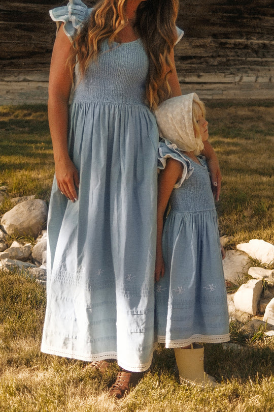 A woman and a child stand together on the grass outdoors, both in matching light blue dresses with smocked bodices. The child wears the Ivy City Co Mini Hattie Dress in Embroidered Chambray, featuring delicate flutter sleeves and paired with a white bonnet, as she leans against the woman.