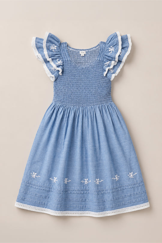 The Mini Hattie Dress in Embroidered Chambray by Ivy City Co is shown laid flat on a neutral background, featuring a light blue smocked bodice, delicate floral embroidery on the skirt, and flutter sleeves trimmed with white lace.