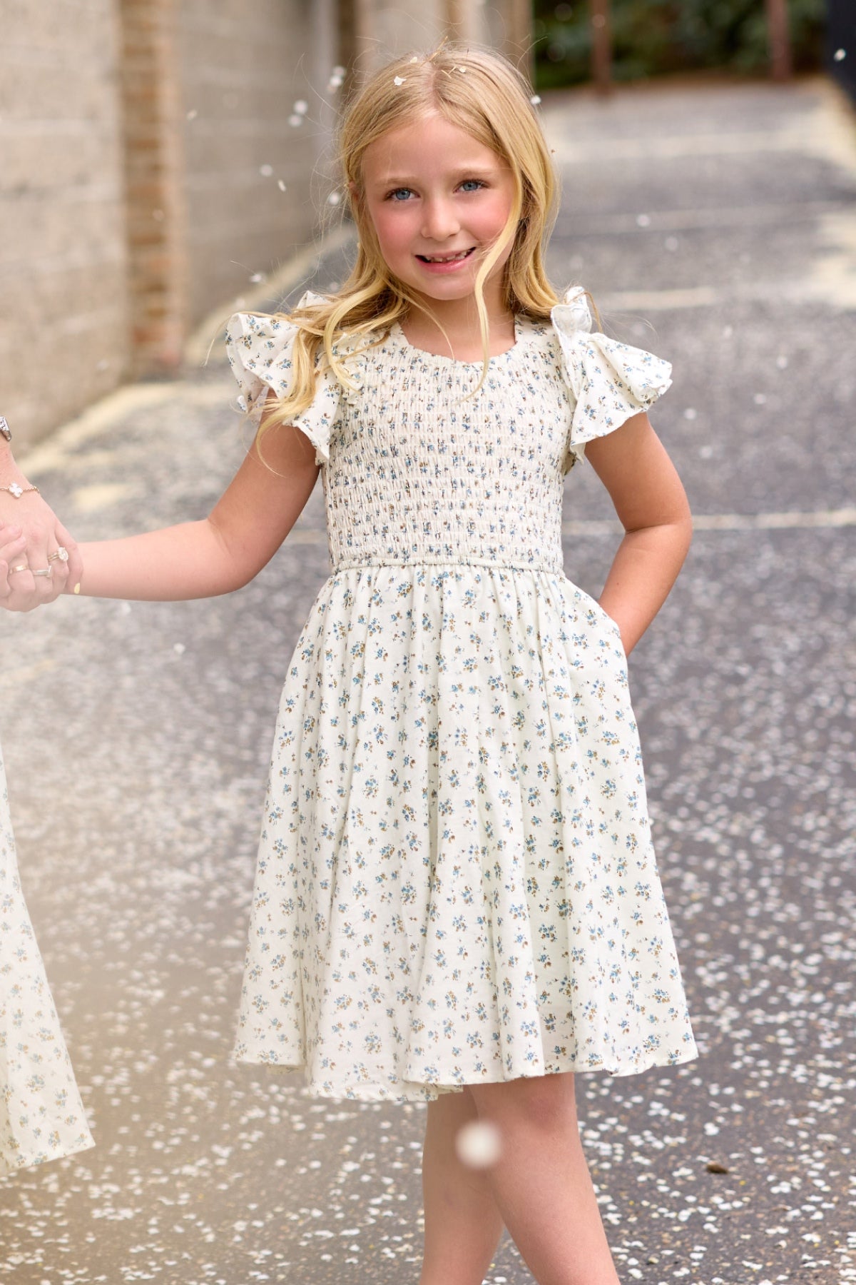 A young blonde girl stands outdoors, smiling and holding someone's hand as petals float around her, wearing the Ivy City Co Mini Hattie Dress in Madeline Blue.