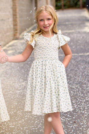 A young blonde girl stands outdoors, smiling and holding someone's hand as petals float around her, wearing the Ivy City Co Mini Hattie Dress in Madeline Blue.