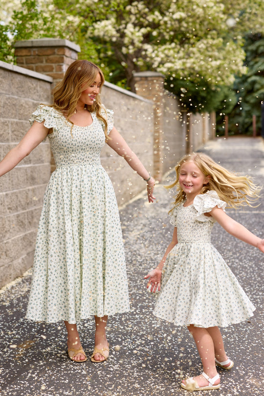A woman and a young girl, both in Ivy City Co’s Mini Hattie Dress in Madeline Blue, smile and play outside among falling flower petals. They show off the charming mommy-and-me look near a stone wall and blooming trees.
