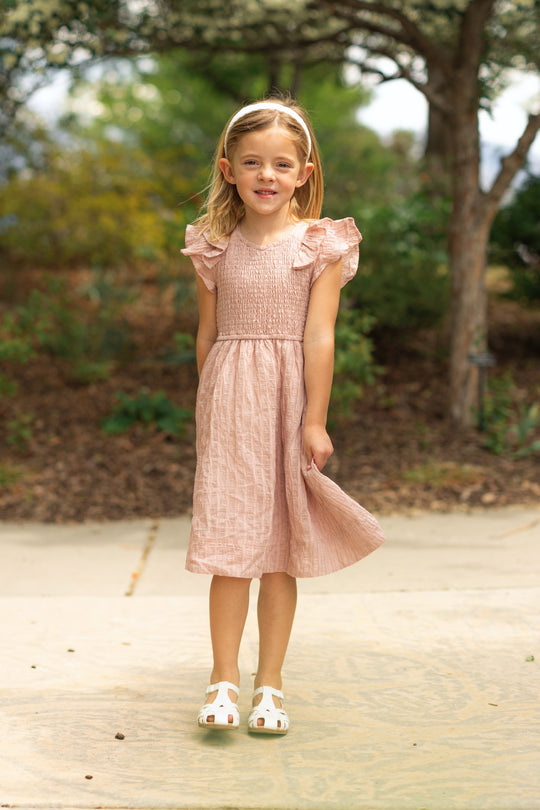 A young girl stands outside smiling in the Ivy City Co Mini Hattie Dress in Pink Stripes with ruffled sleeves and white sandals. Her shoulder-length blonde hair is styled with a headband among trees and greenery.