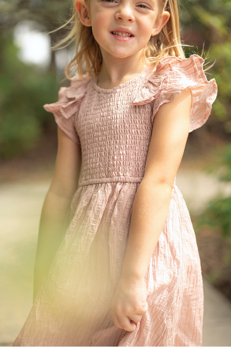 A smiling young girl with light hair wears the Ivy City Co Mini Hattie Dress in Pink Stripes—featuring ruffled shoulders—against a green, outdoor backdrop for a sweet Mom and Me matching moment.