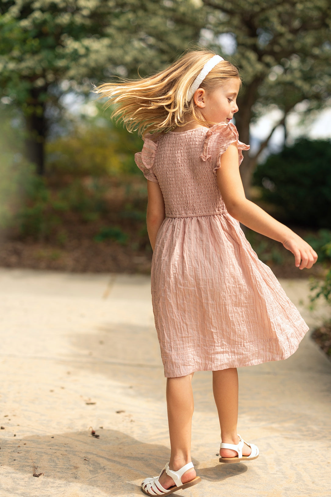 A young girl in the Ivy City Co Mini Hattie Dress in Pink Stripes twirls outdoors on a sunlit path, her girls summer dress flowing as trees and greenery frame the scene.
