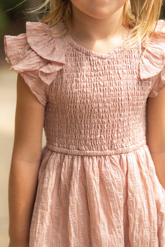 A young child stands outdoors wearing the Ivy City Co Mini Hattie Dress in Pink Stripes—a pale pink girls’ summer dress with a smocked bodice and ruffled sleeves. The child’s face is partially out of frame and slightly blurred.
