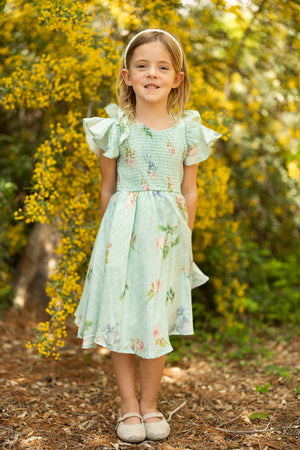 A young girl smiles outdoors on a dirt path, wearing the Ivy City Co Mini Hattie Dress in Roselyn Blue and a matching headband, standing in front of yellow flowering bushes and greenery.
