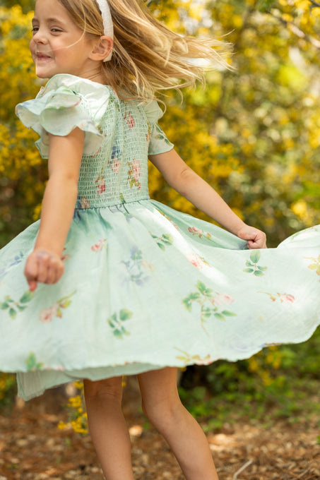 A young girl with blonde hair wearing the Ivy City Co Mini Hattie Dress in Roselyn Blue and a white headband twirls joyfully outdoors, surrounded by yellow flowers and greenery in bright sunlight.