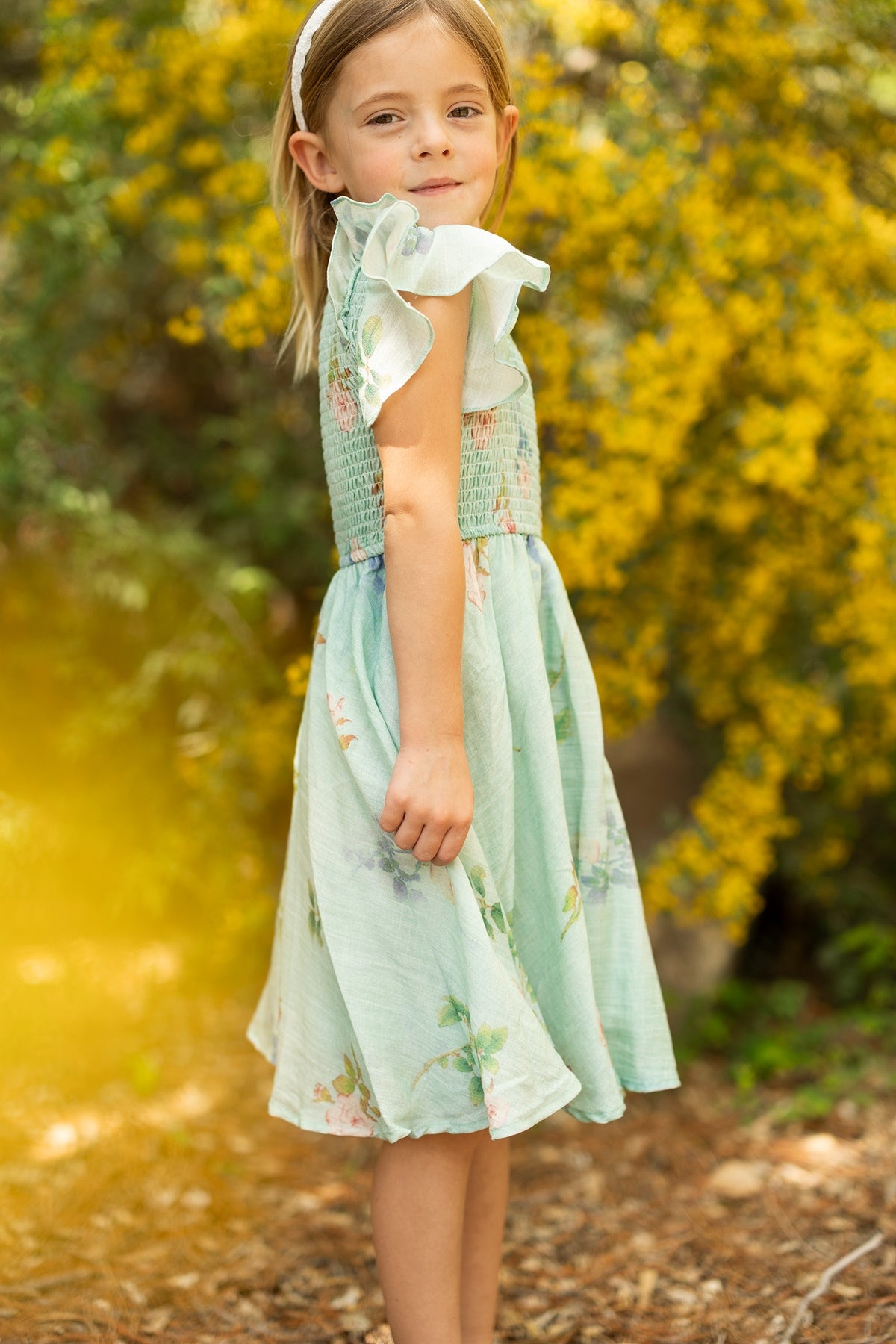 A young girl with light brown hair, wearing the Ivy City Co Mini Hattie Dress in Roselyn Blue and a white headband, stands outdoors near yellow flowering bushes, looking at the camera with a slight smile.
