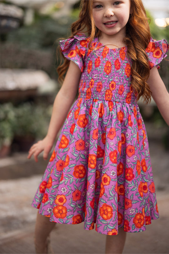 A young girl with long red hair smiles outdoors, wearing the Mini Hattie Dress in Thistle Flower Poppy Woodblock by Ivy City Co—a girls' cotton midi dress with puffy sleeves and a vibrant floral print on a purple background.