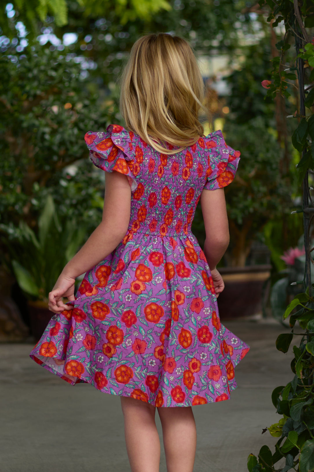 A young girl with blonde hair stands outdoors, facing away, wearing the Ivy City Co Mini Hattie Dress in Thistle Flower Poppy Woodblock. Green plants and trees surround her in the background.