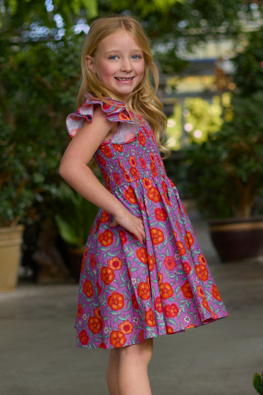 A young girl with long blonde hair smiles in a garden, wearing Ivy City Co’s Mini Hattie Dress in Thistle Flower Poppy Woodblock— a purple floral dress with red and orange flowers and a smocked bodice, standing sideways with one hand in her pocket.