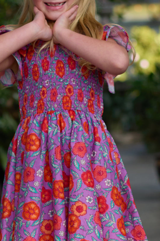 A young child wears the Ivy City Co Mini Hattie Dress in Thistle Flower Poppy Woodblock—a purple floral cotton midi with ruffled sleeves and a smocked bodice—smiling outdoors with hands under their chin.