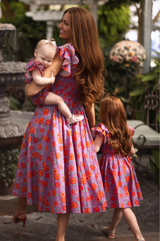 A woman with long brown hair wears the Ivy City Co Mini Hattie Dress in Thistle Flower Poppy Woodblock, holding a baby in a matching floral outfit while a young girl in similar attire walks beside them through a lush garden.