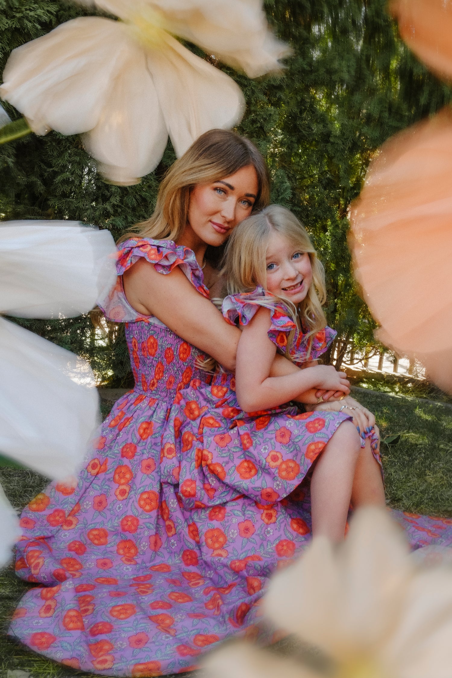 A woman and young girl smile as they sit outdoors, both wearing Ivy City Co's Mini Hattie Dress in Thistle Flower Poppy Woodblock. Surrounded by soft-focus flowers, they embrace in matching purple floral dresses with smocked bodices.
