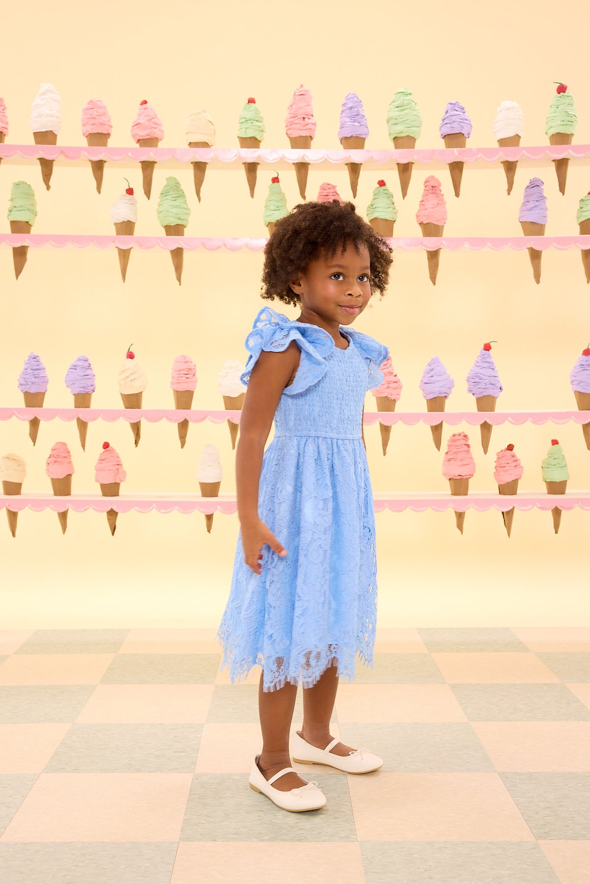 Child in a blue dress standing in front of an ice cream cone wall.