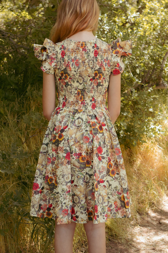 A girl with long light brown hair stands outdoors, back to the camera, in Ivy City Co’s Mini Hattie Dress in Rose Vine—100% cotton, gathered bodice, ruffled shoulders—surrounded by green foliage.