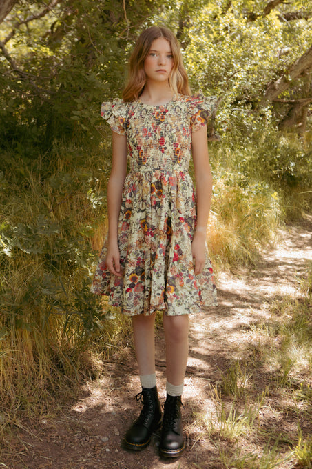 A young girl stands on a wooded path wearing the Ivy City Co Mini Hattie Dress in Rose Vine, featuring 100% cotton with ruffled sleeves, paired with white socks and black boots as sunlight shines through the trees.