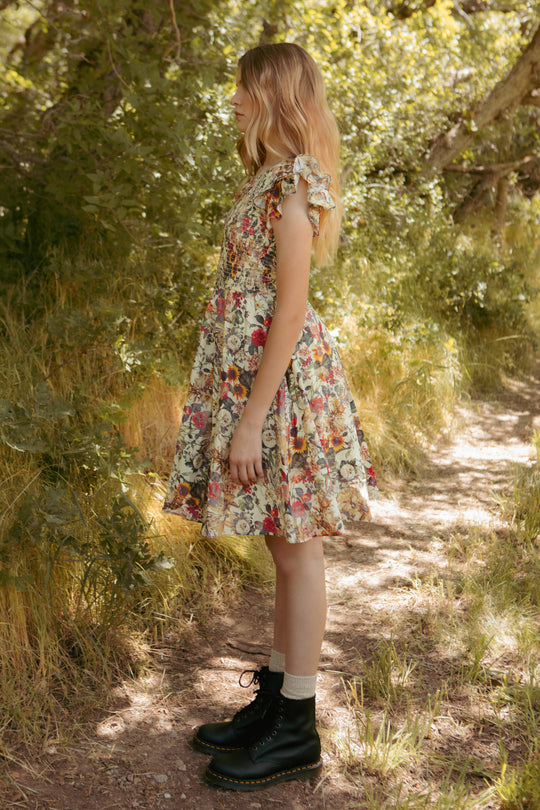 A young woman stands in profile on a forest path, wearing Ivy City Co’s Mini Hattie Dress in Rose Vine print with ruffled sleeves, light socks, and black lace-up boots. She is surrounded by lush green foliage.