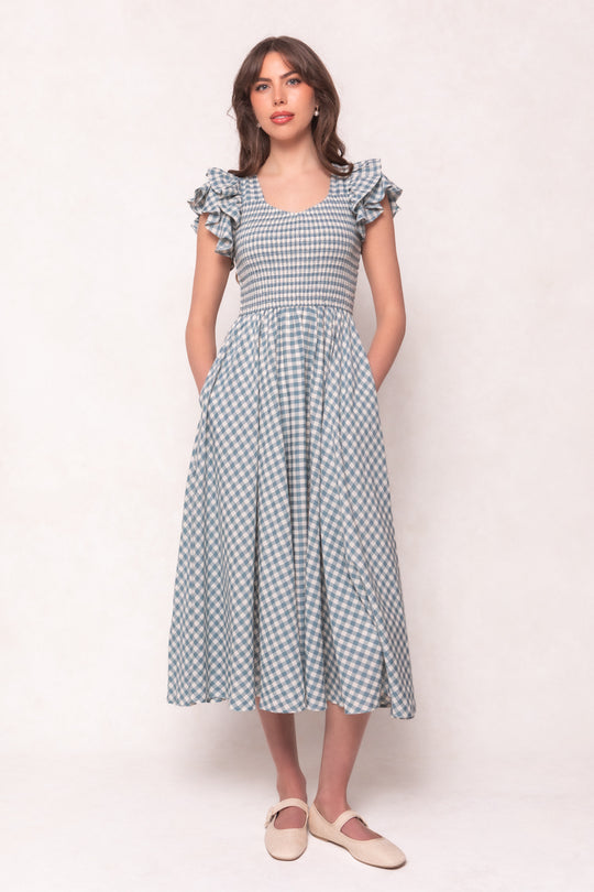 A woman stands against a light background wearing the Hattie Dress in Dusty Teal Gingham by Ivy City Co, a maternity-friendly cotton midi dress with ruffled sleeves, fitted bodice, pockets, and cream-colored flats.