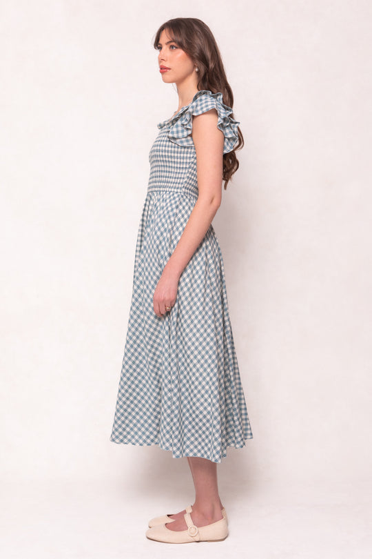 A woman with long brown hair poses in profile against a plain background, wearing the Ivy City Co Hattie Dress in Dusty Teal Gingham with ruffled sleeves and beige flats—a stylish, maternity-friendly cotton midi dress.