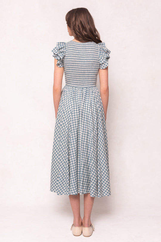 A woman wears the Ivy City Co Hattie Dress in Dusty Teal Gingham—a cotton, maternity-friendly midi dress with ruffled short sleeves and a fitted waist—paired with beige shoes against a light background.