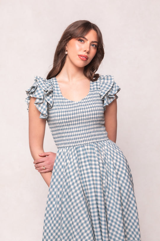 A woman with long brown hair wears the Ivy City Co Hattie Dress in Dusty Teal Gingham, featuring ruffled sleeves. She stands against a light background, smiling slightly at the camera with her hand resting on her opposite arm.