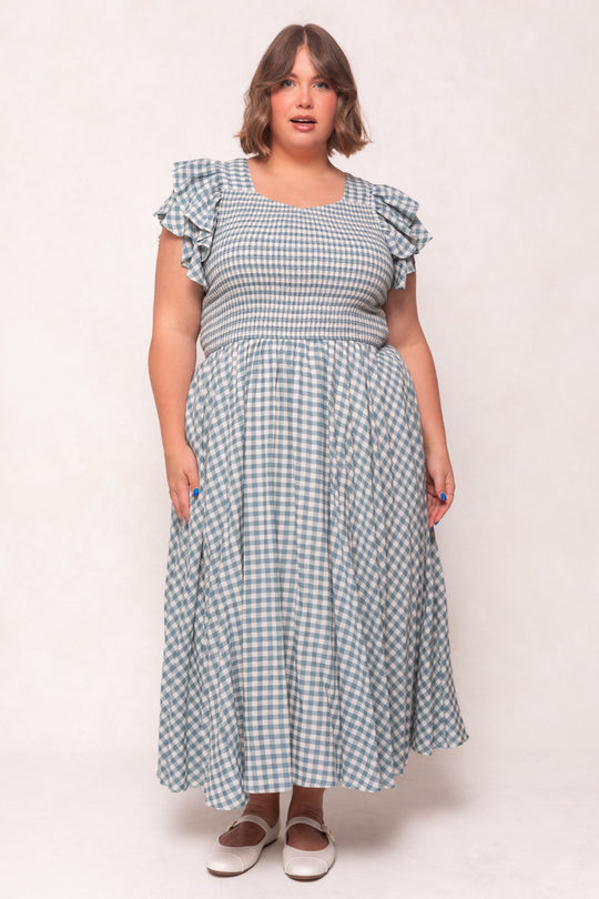 A woman with short brown hair wears the Ivy City Co Hattie Dress in Dusty Teal Gingham, featuring ruffled sleeves, and white Mary Jane shoes, standing against a plain light background.