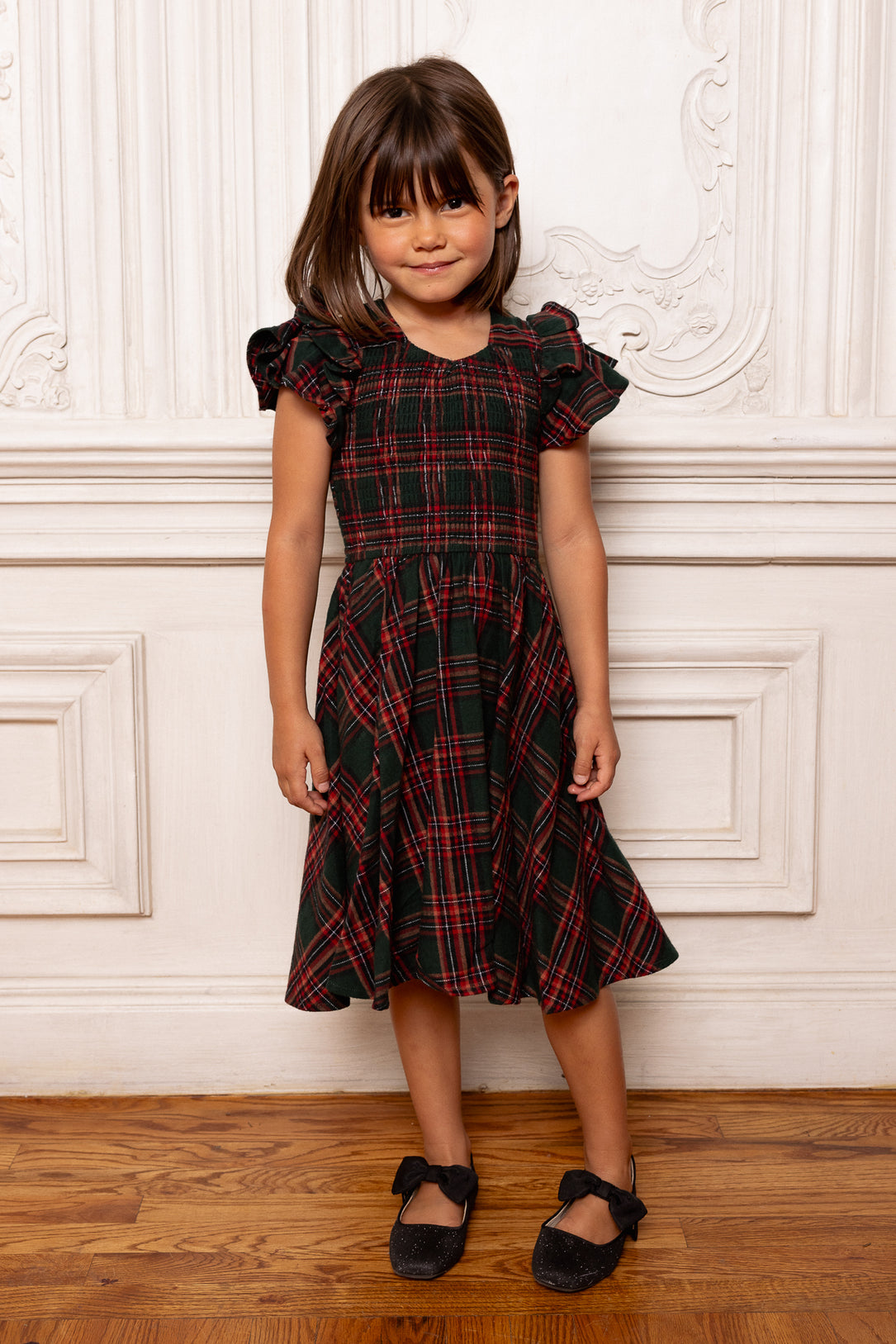 Young girl wearing a black and red plaid dress standing against a white wall.